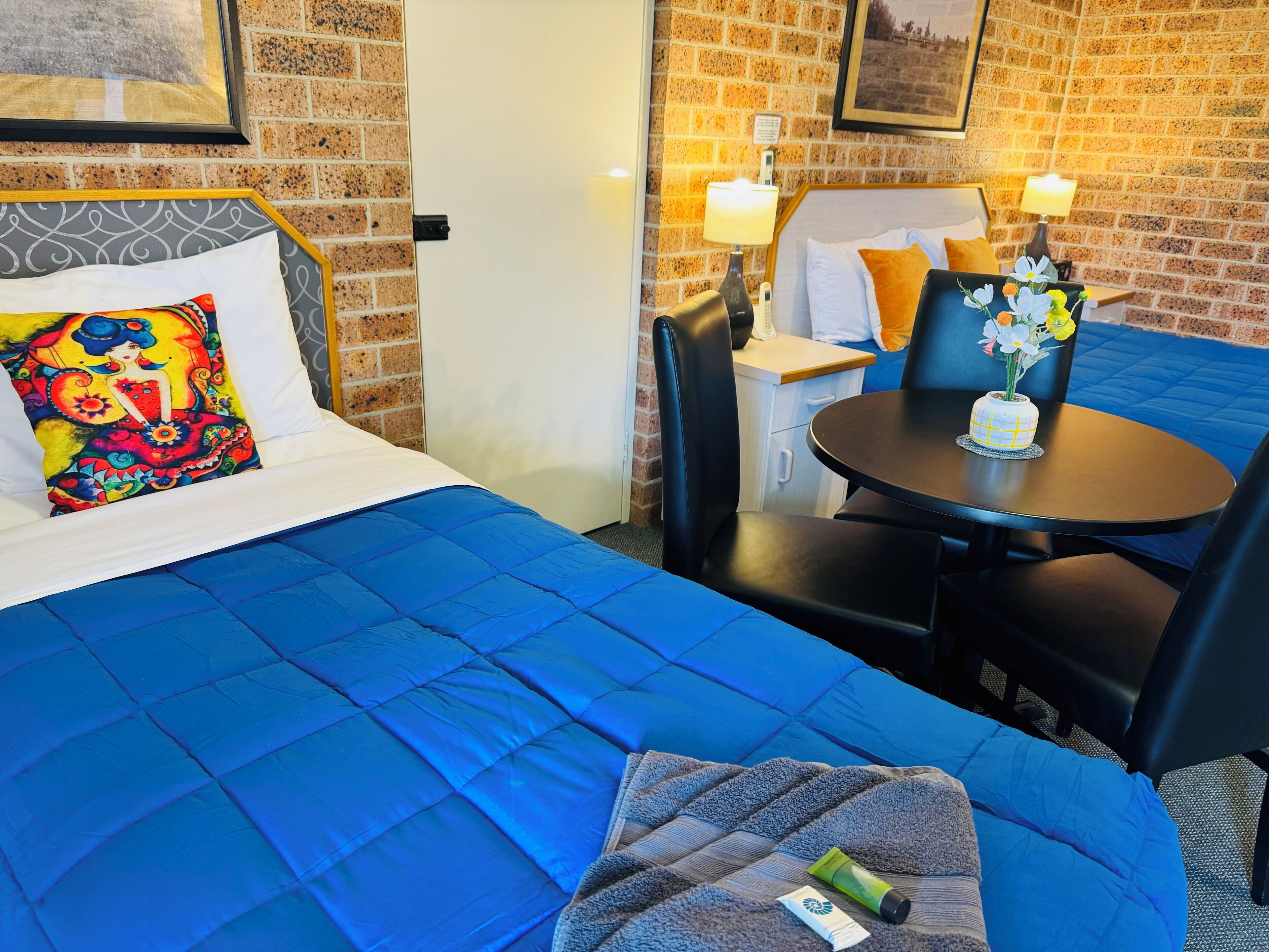 A hotel room with a bed, a small table with chairs, a vase of flowers, and a brick wall.
