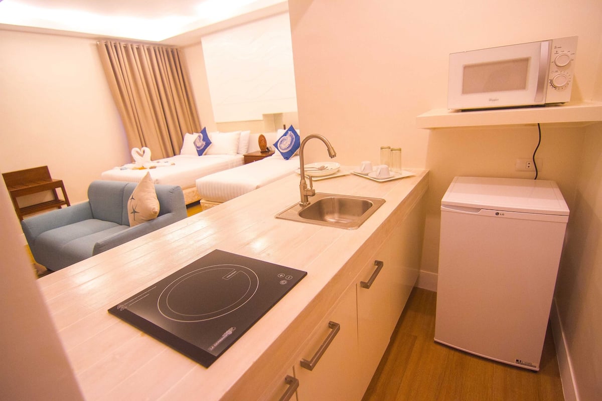 A compact hotel room with a kitchenette, a bed with blue and white bedding, a small seating area, and a microwave on a shelf.