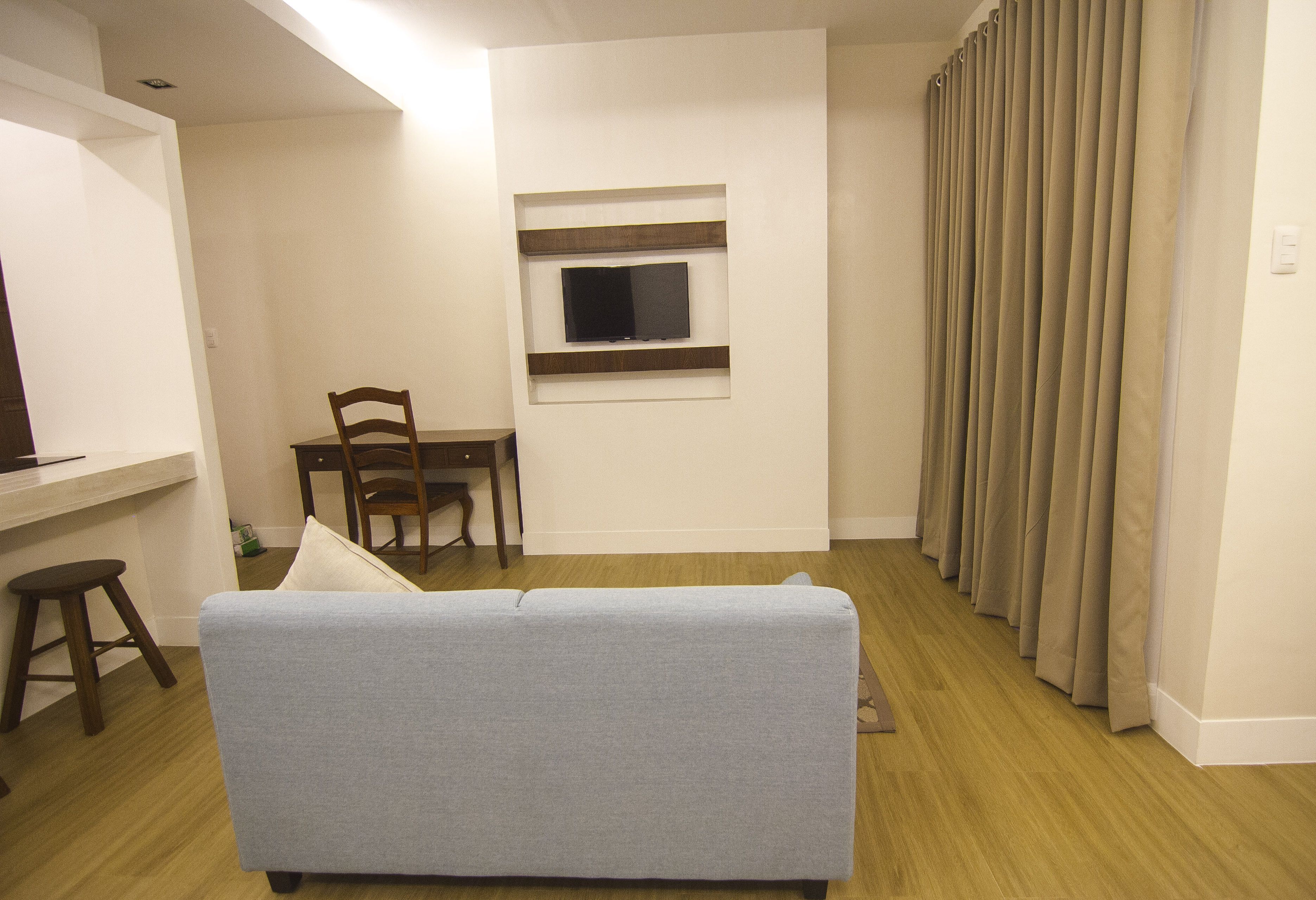 A living room with a grey sofa, a TV mounted on the wall, a dining area with a table and chairs, and a window with curtains.