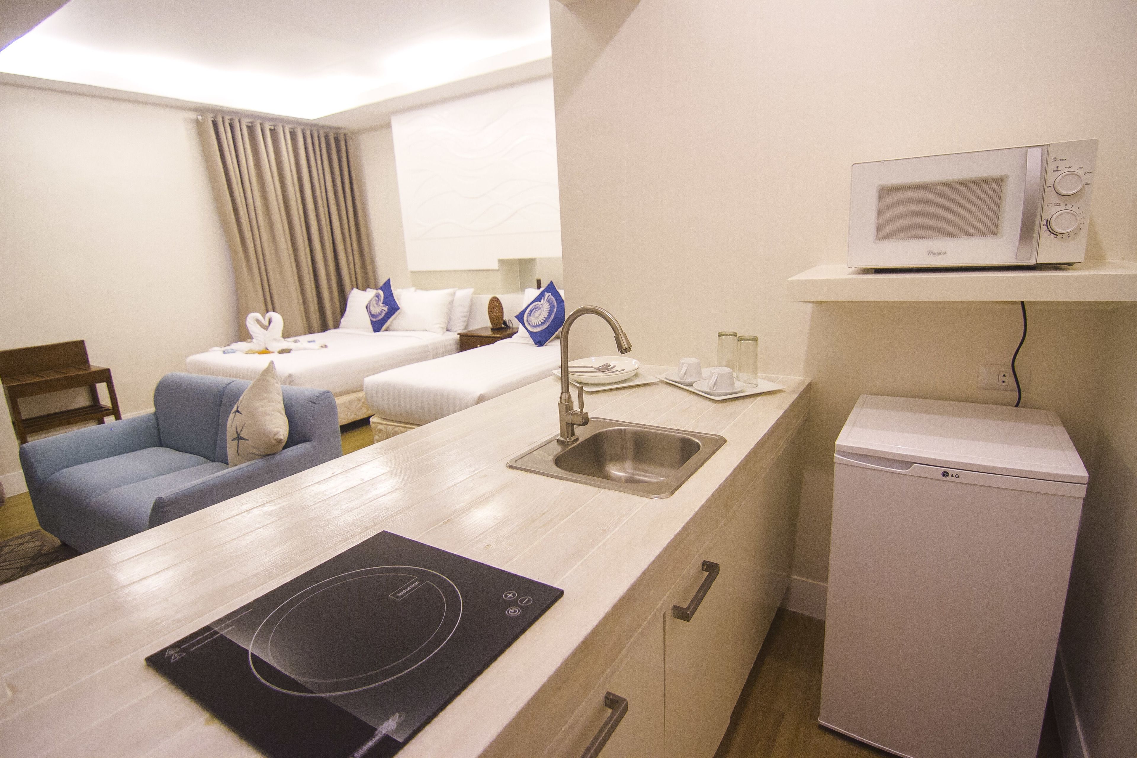 Camera d'albergo compatta con angolo cottura, forno a microonde, frigorifero, lavello e un letto con una sedia blu e cuscini decorativi.