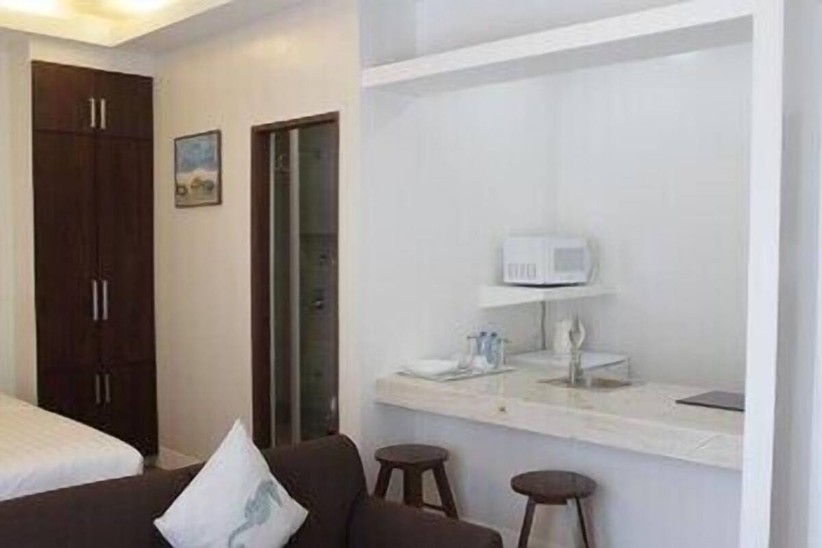 A hotel room with a bed, a dark wooden wardrobe, a small table with a white lamp, and a wall-mounted microwave.