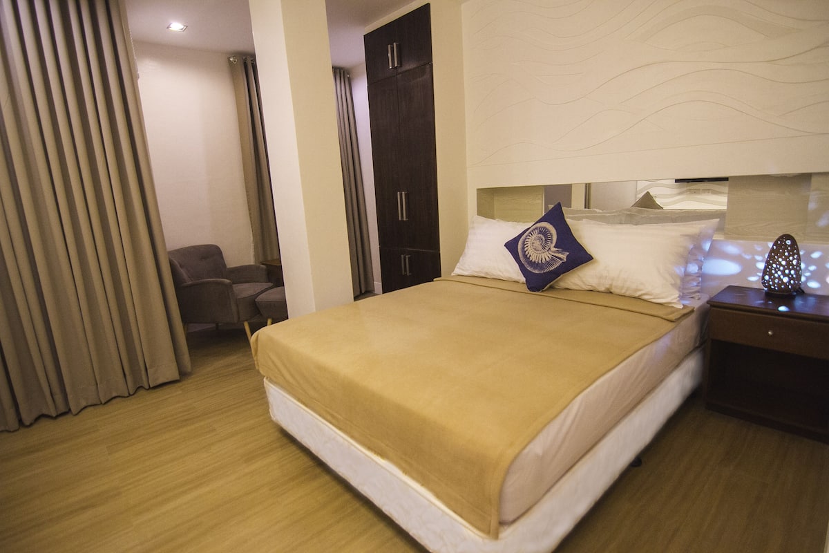 A hotel room with a large bed, a chair, a wardrobe, and a nightstand.