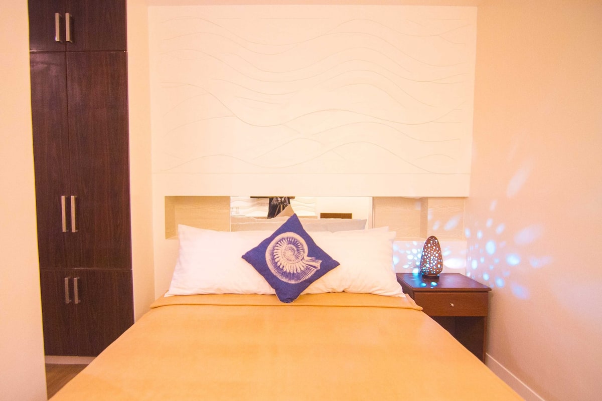 A hotel room with a bed, a nightstand, a decorative pillow, and a wall-mounted light fixture.