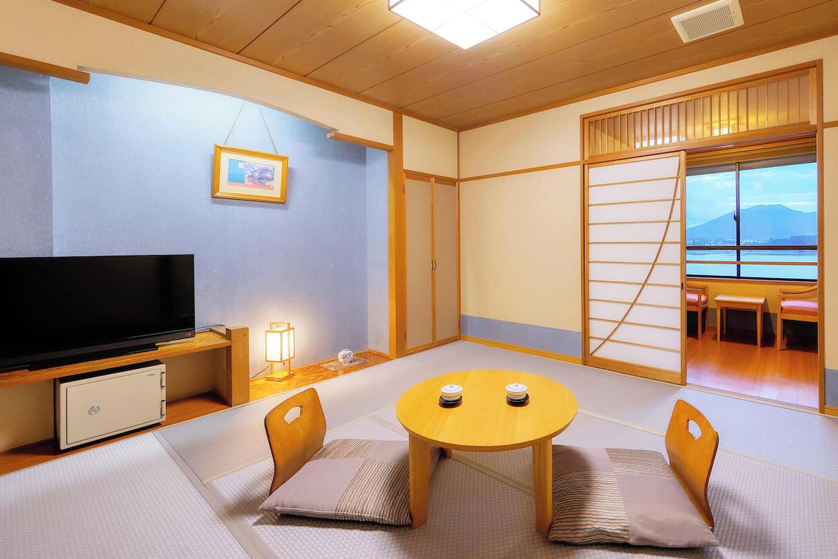 Une pièce de style japonais traditionnel, avec un sol recouvert de tatamis, une télévision, un petit meuble, une table avec des coussins et une porte coulissante donnant sur un balcon.