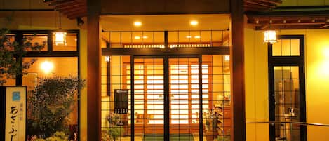 Entrada de un edificio tradicional japonés con un portón de madera, plantas en macetas y un felpudo verde en la entrada.