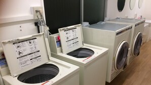 A row of front-loading washing machines with instructional labels, a water inlet, and a wooden floor. - Glocal Nagoya Backpackers Hostel (Nagoya)