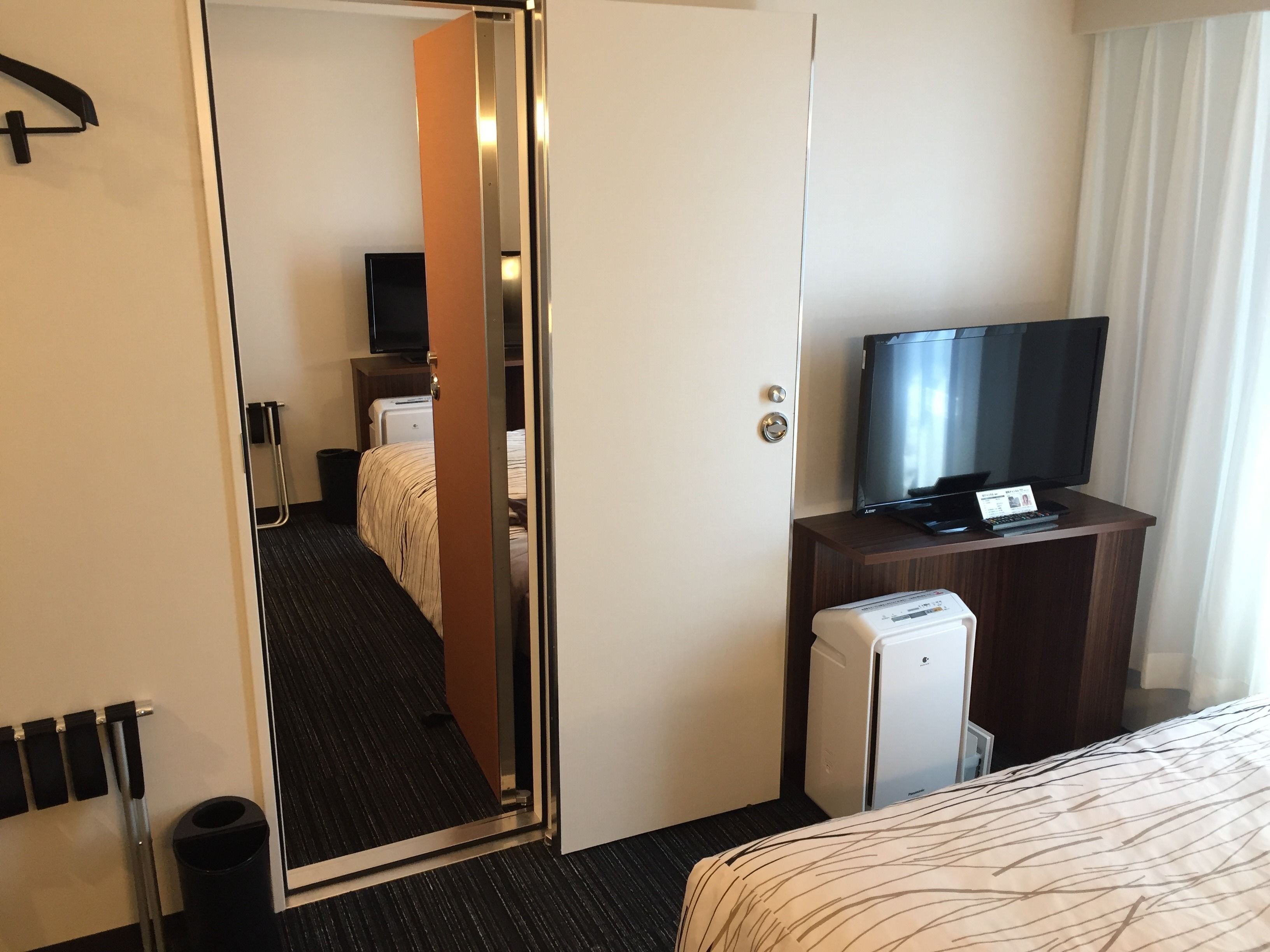 A hotel room with a bed, a television on a wooden dresser, a minibar, and a mirror reflecting the room.