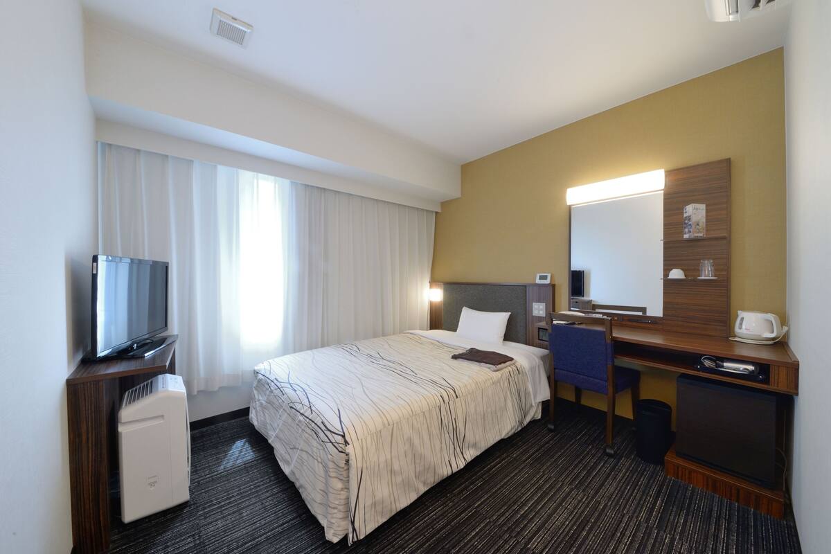A hotel room with a bed, a desk with a chair, a television, a mini-fridge, and a mirror.