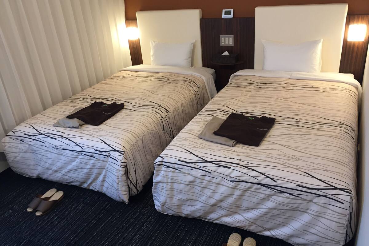 A hotel room with two beds, each with a striped bedspread and white pillows. There is a small table between the beds with a tissue box on it.