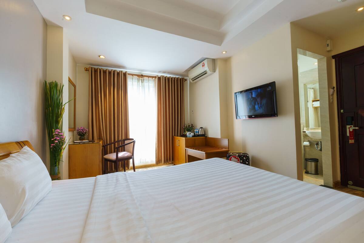 A hotel room with a large bed, a desk with a chair, a television, and an air conditioning unit.