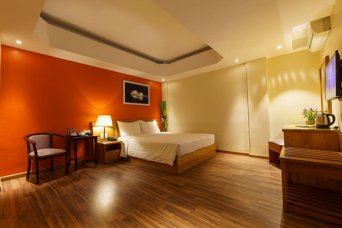 A hotel room with a wooden floor, a bed, a desk with a lamp, a chair, and a TV.