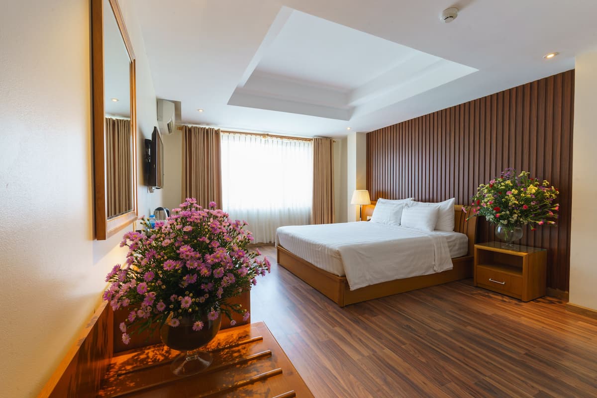 A hotel room with a large bed, a wooden floor, a mirror, a TV, and a vase of flowers.