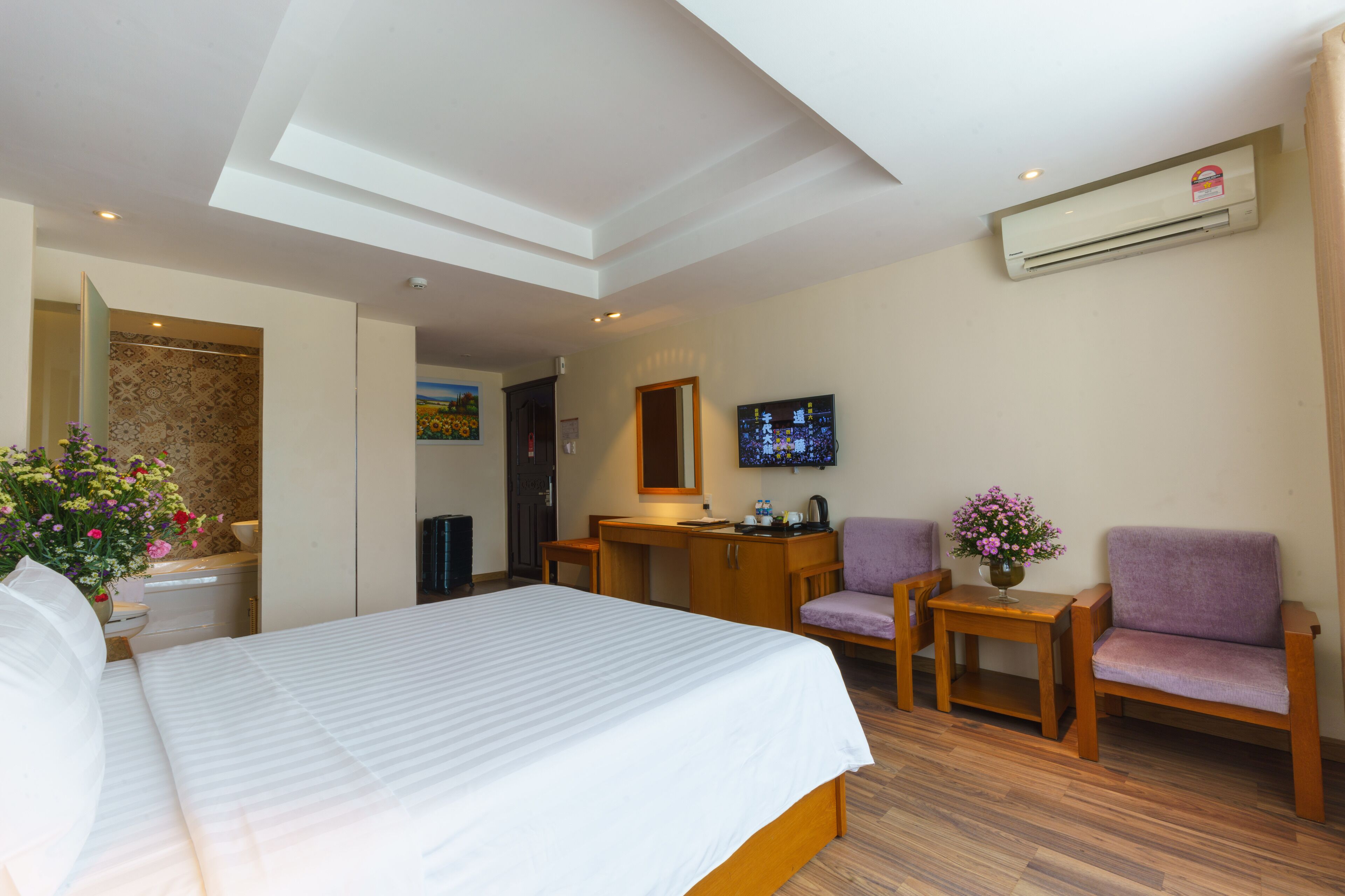 A hotel room with a bed, a desk, a chair, a small table with flowers, and an air conditioning unit.