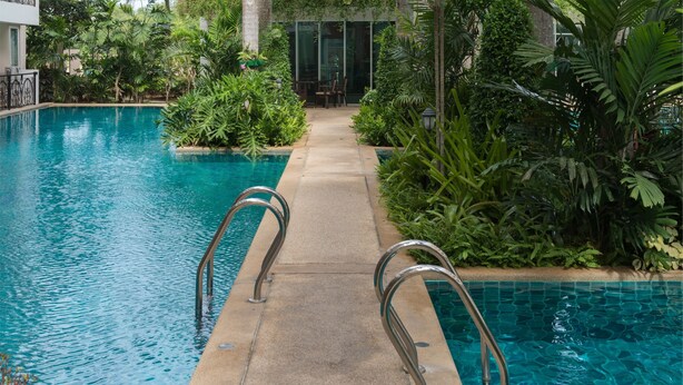Une piscine dotée de rampes métalliques, entourée d’une végétation luxuriante et bordée d’un chemin menant à un bâtiment.