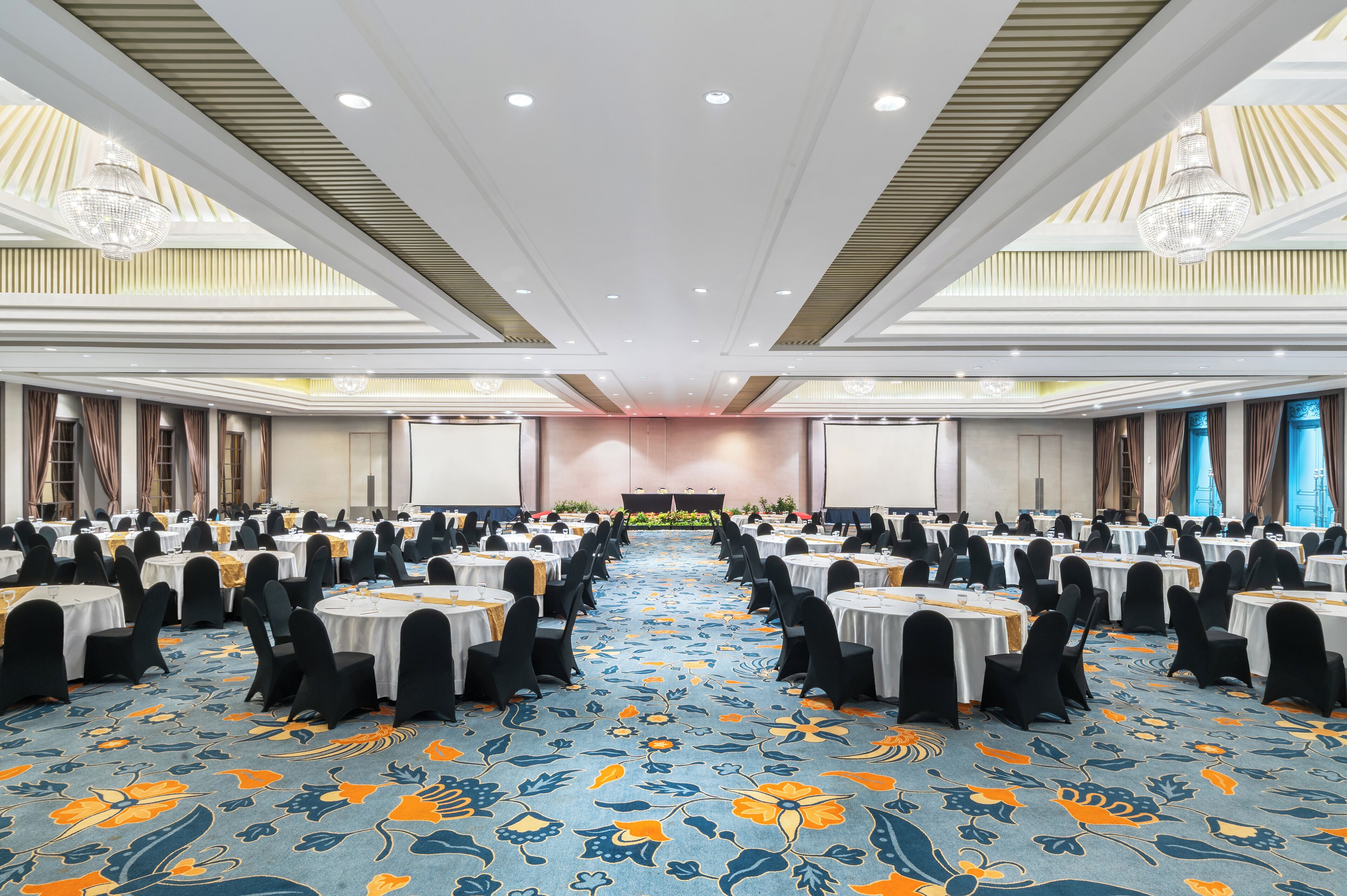 A banquet hall with rows of round tables, black chairs, and a floral carpet.