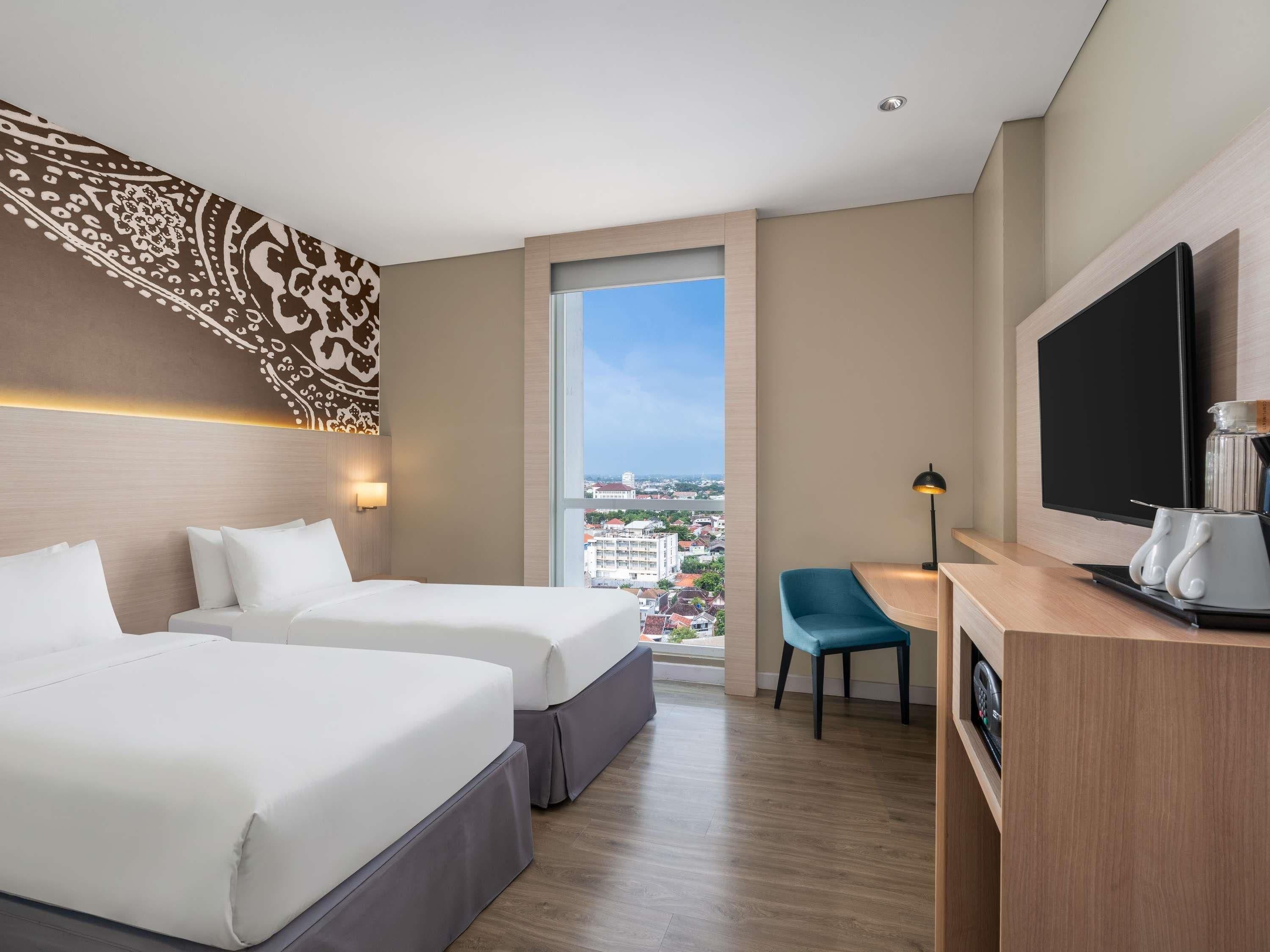 Une chambre d’hôtel avec deux lits, un bureau avec une télévision, une chaise et une grande fenêtre donnant sur la ville.