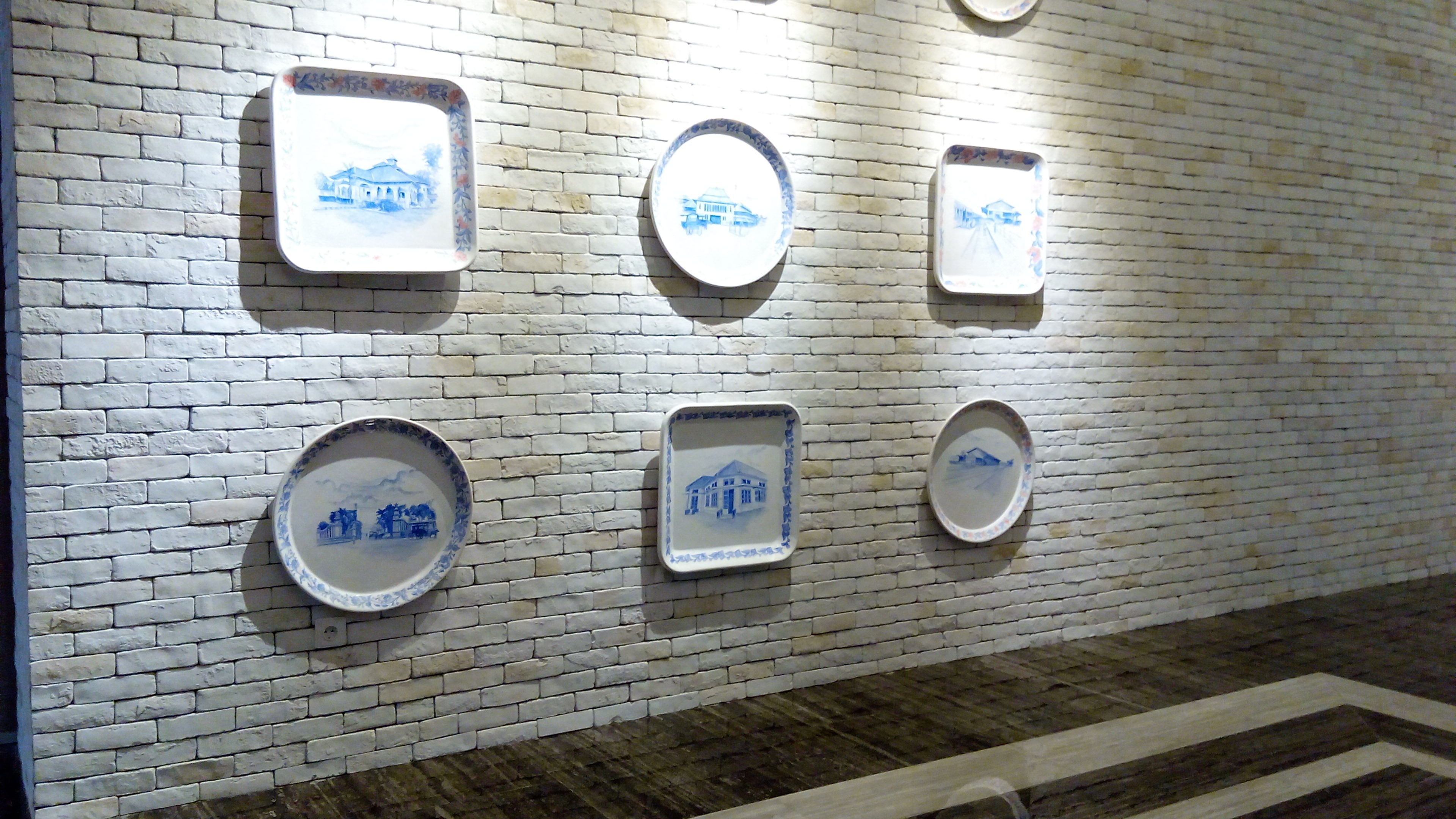 Un mur orné de nombreuses assiettes en céramique, de forme carrée ou ronde, décorées de motifs bleus, et fixées sur un mur en briques blanches.