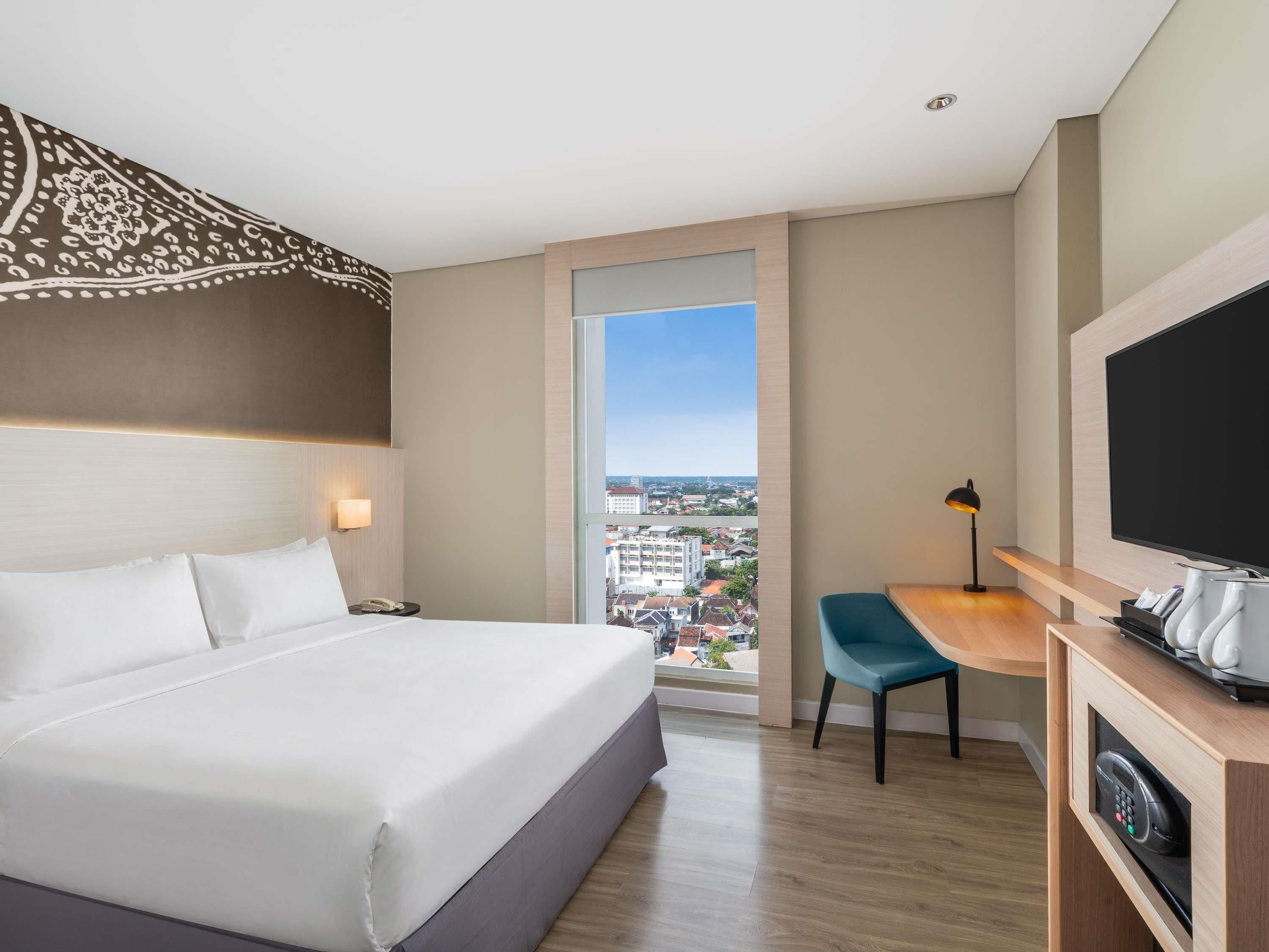 A hotel room with a bed, a desk, a television, and a view of the city.