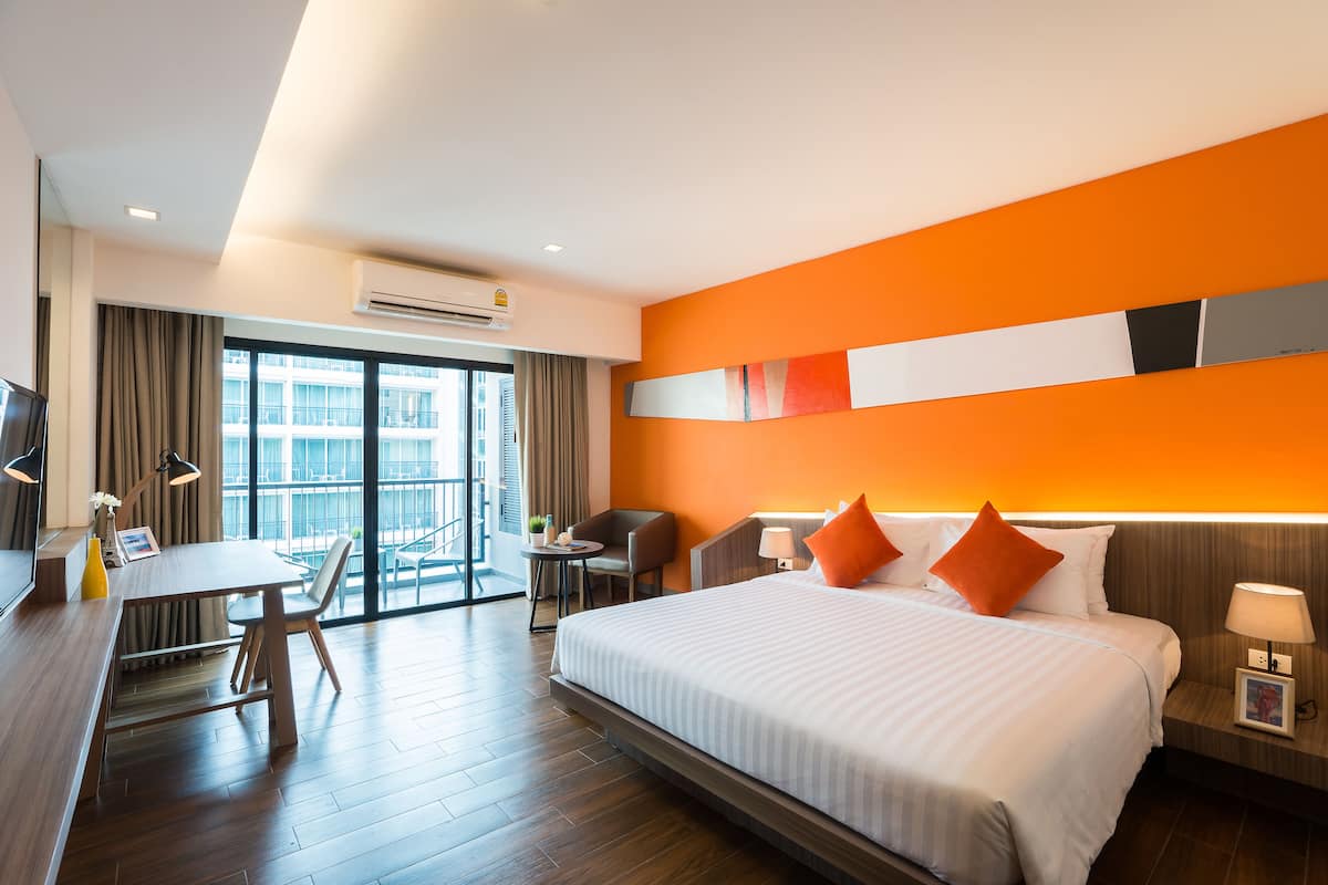 A hotel room with a large bed, a desk, and a chair. There is a window with curtains, an air conditioning unit, and a view of a building outside.