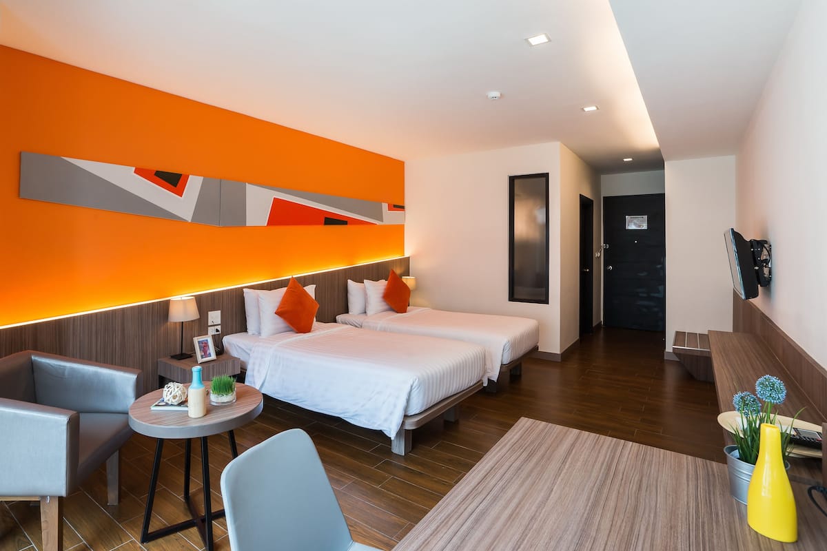 A modern hotel room with two beds, a seating area, and a bright orange accent wall.