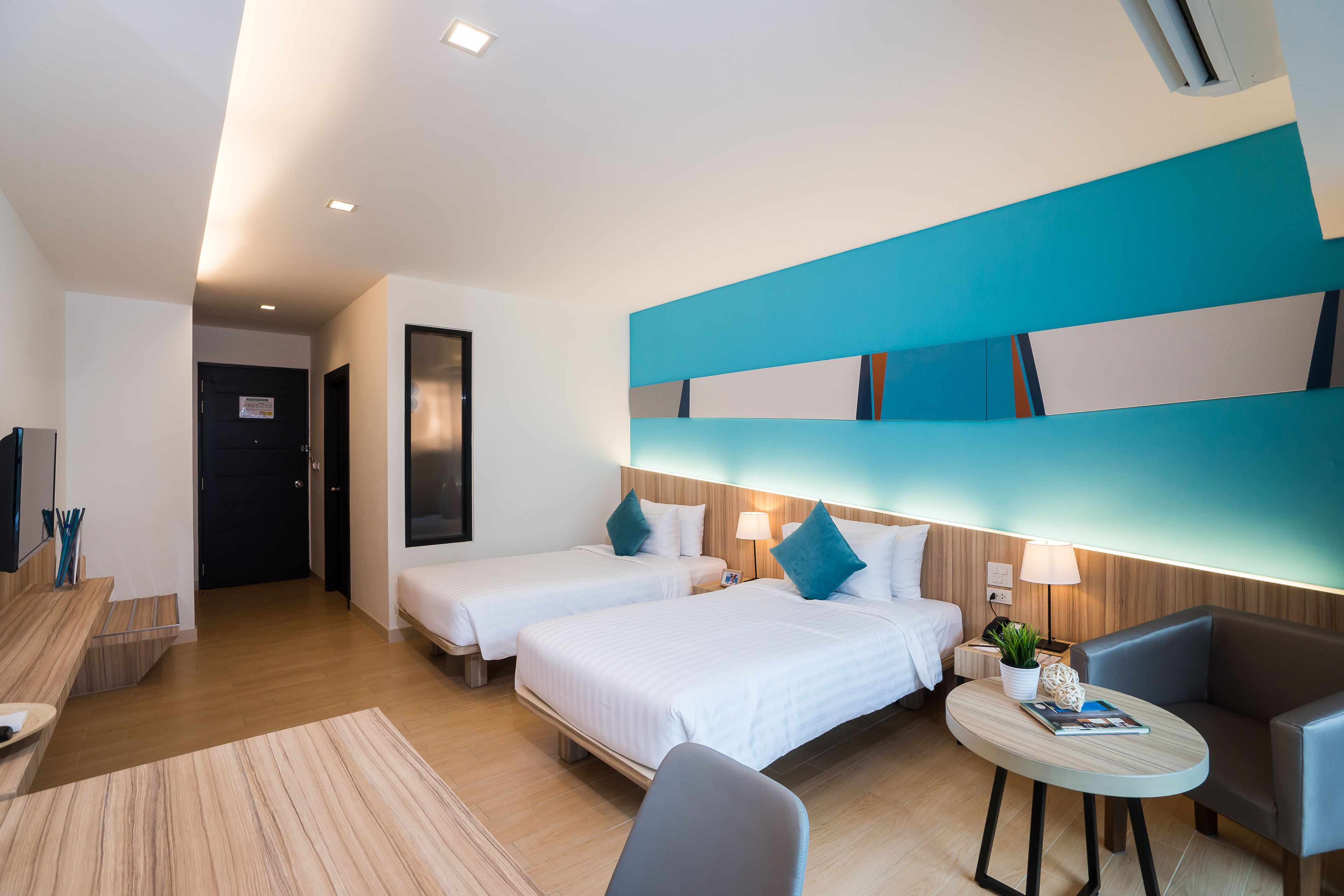 A hotel room with two beds, a desk, a chair, a small table with a lamp, and a wall with a geometric design.