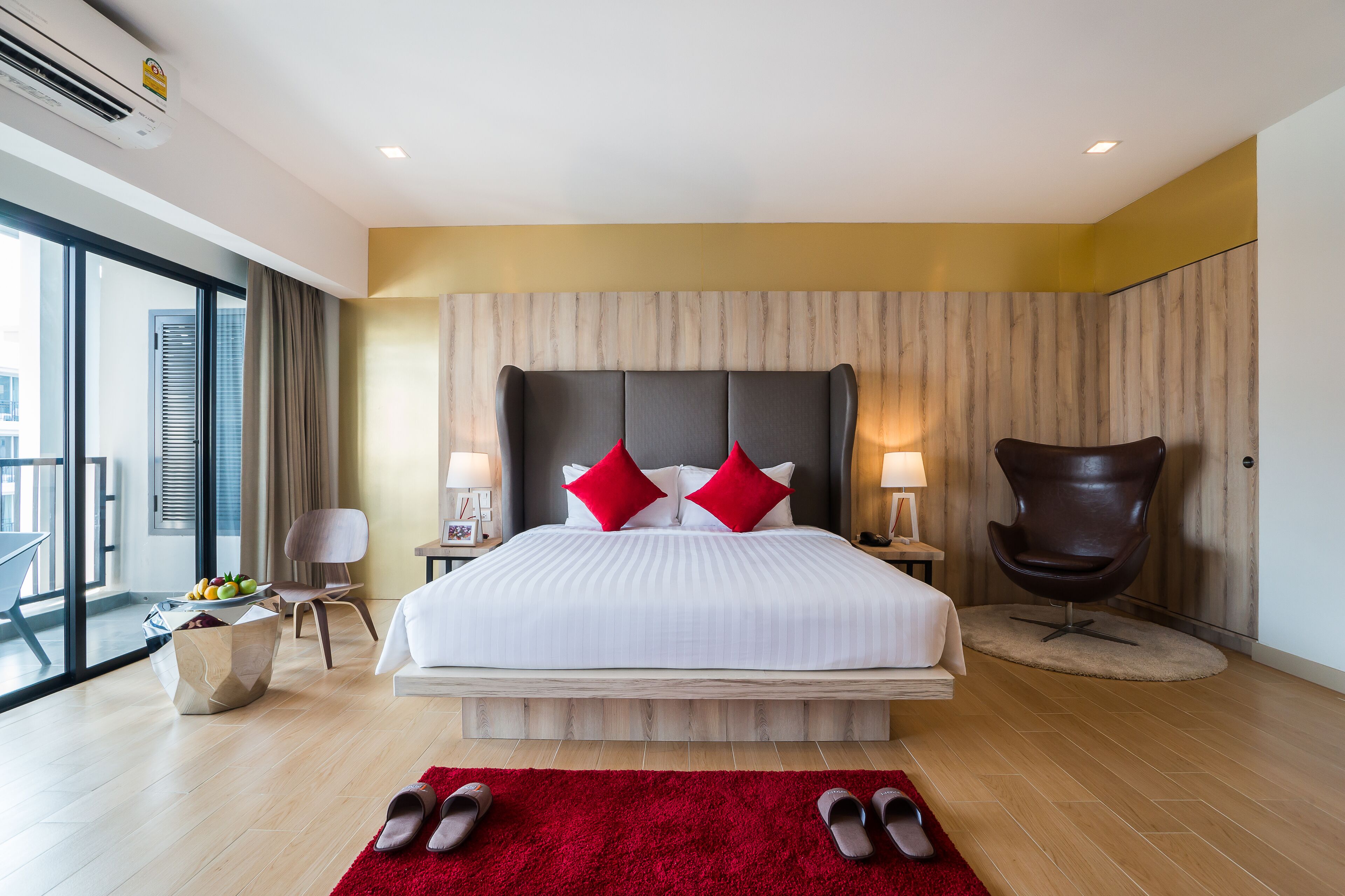 A modern hotel room with a large bed, a red rug, and a view of the city through large windows.