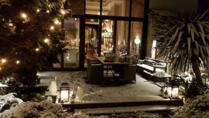 Un espace extérieur enneigé, avec une table et des chaises, entouré de plantes en pot et illuminé par des guirlandes lumineuses.