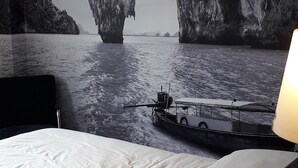 Une chambre d’hôtel avec un lit, une grande fresque murale représentant un paysage côtier, une lampe suspendue et une table de chevet.