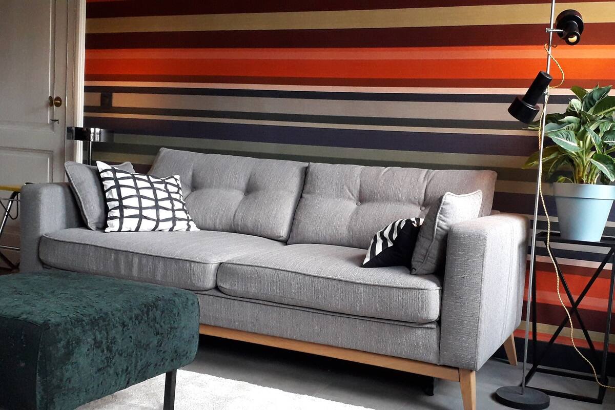 A grey sofa with multiple cushions, a green ottoman, a floor lamp, and a potted plant against a striped wall.