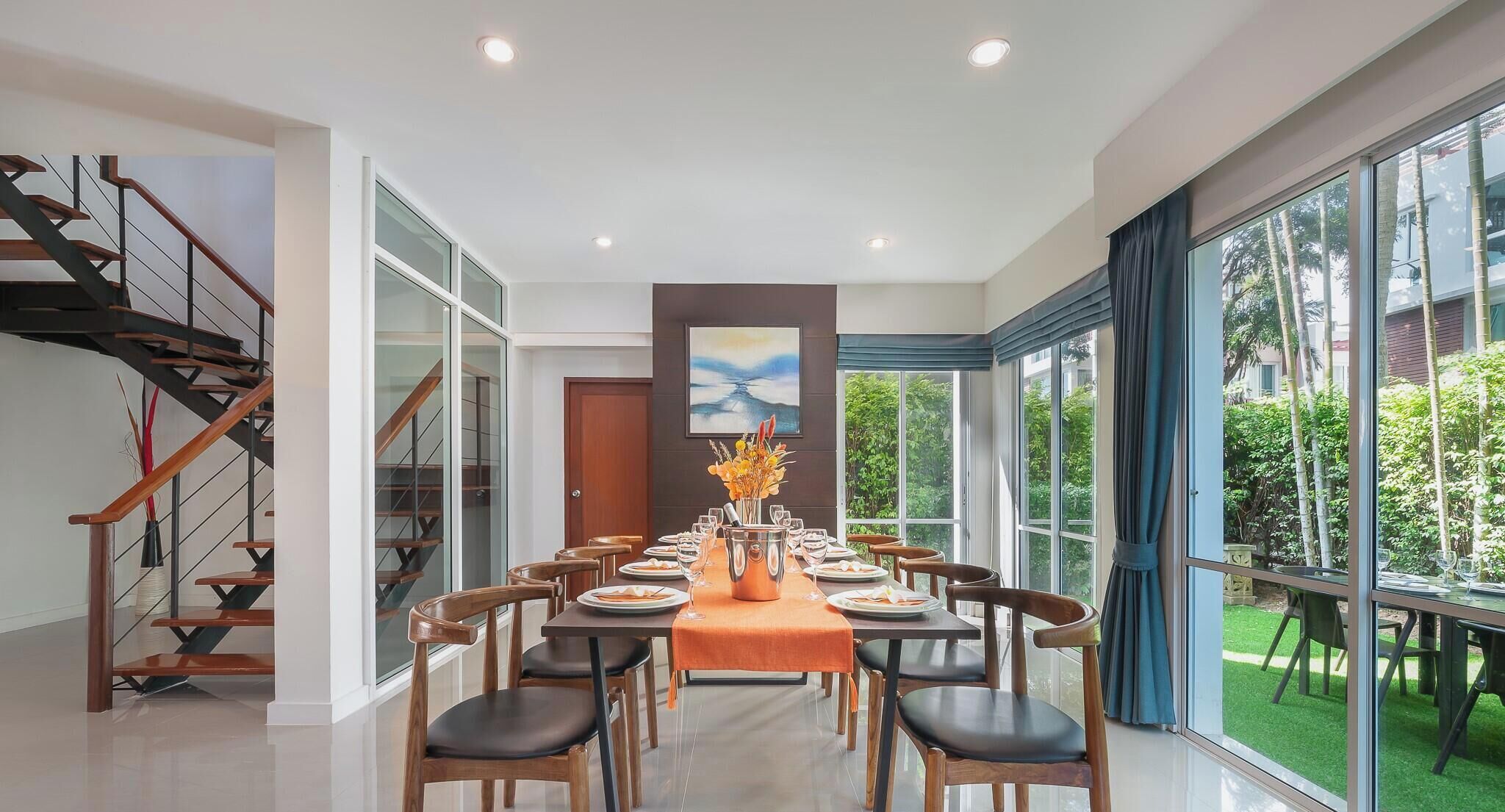 A dining area with a long table set for a meal, surrounded by chairs, and a staircase on the left.
