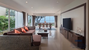 Ein modernes Wohnzimmer mit Sofa, Couchtisch und Fernseher, großen Fenstern sowie einem Essbereich mit Stühlen und Tisch.