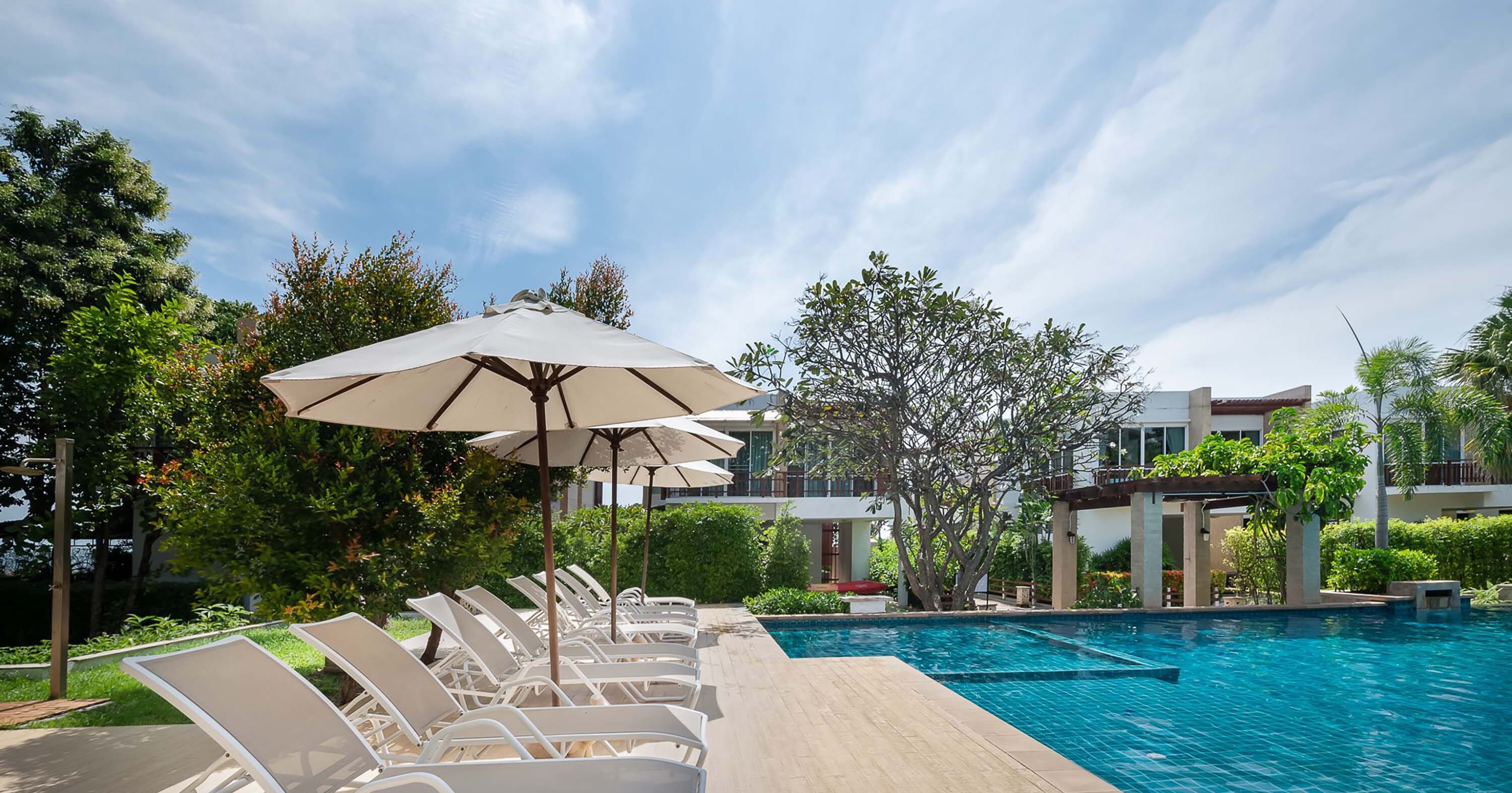 Una piscina con sillas de descanso y sombrillas, rodeada de vegetación y edificios modernos.
