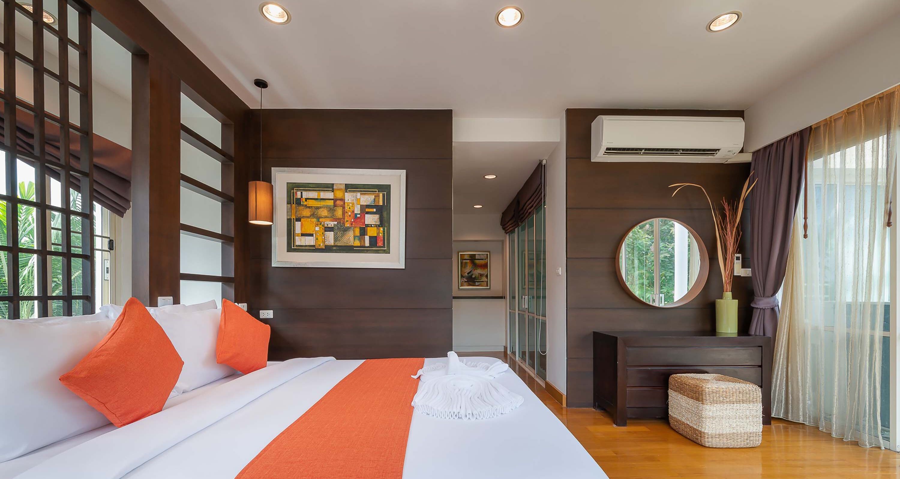 A modern hotel room with a large bed, orange pillows, a framed artwork, an air conditioning unit, a round mirror, and a view of greenery through the window.
