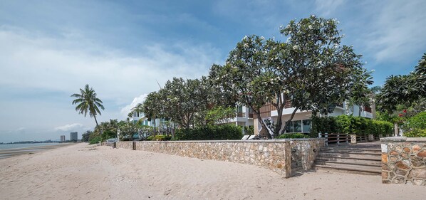 A beachfront property with a stone wall, palm trees, and a building with multiple balconies. - Simantra Private Villas (Cha-am)