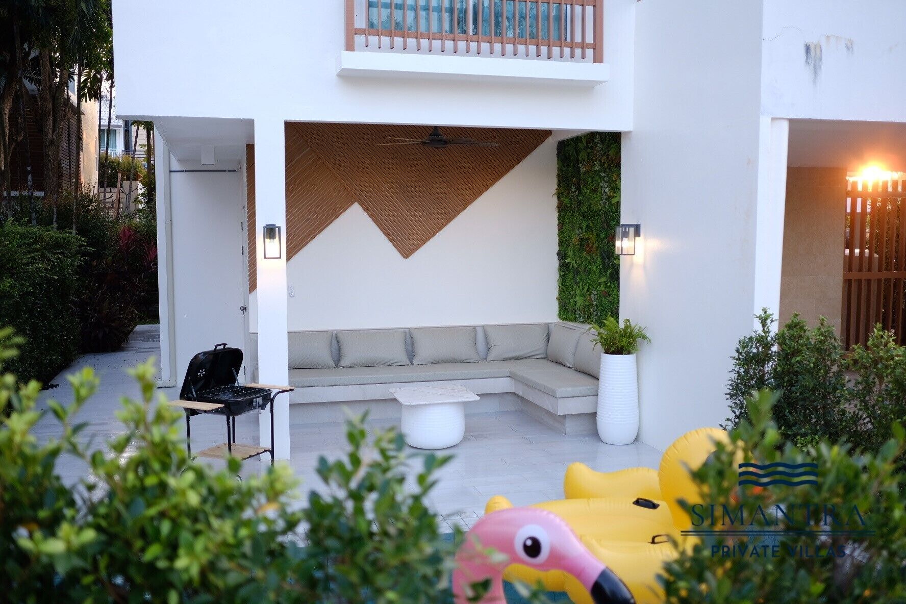A modern outdoor seating area with a sofa and a small table, surrounded by greenery and a decorative flamingo statue.