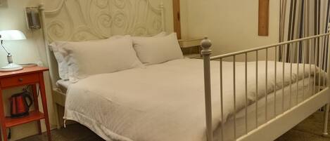 A single-bed room with a white metal headboard, a bedside table with a lamp, and a red bag on a shelf.