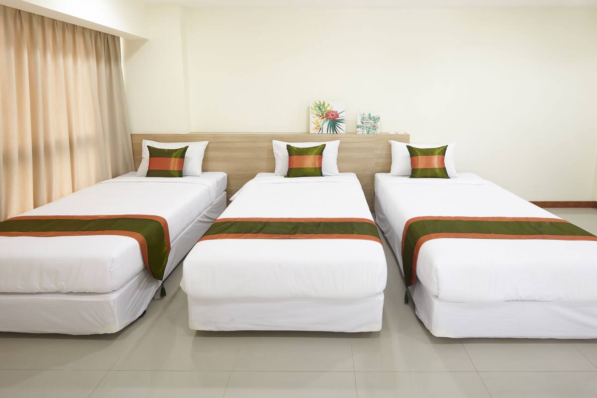 Ein Hotelzimmer mit drei Einzelbetten, jedes mit weißer Bettwäsche und grünen und orangefarbenen Akzenten. Auf jedem Bett liegen zwei Zierkissen. Das Zimmer verfügt über ein hölzernes Kopfteil, beige Vorhänge und ein kleines Regal mit zwei Dekorationselementen.