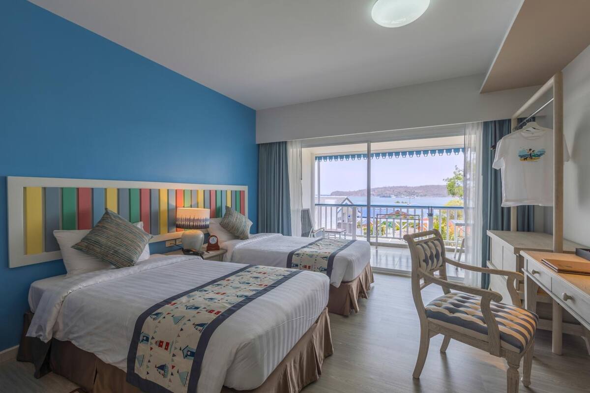 A hotel room with two beds, a desk, a chair, and a view of the sea.