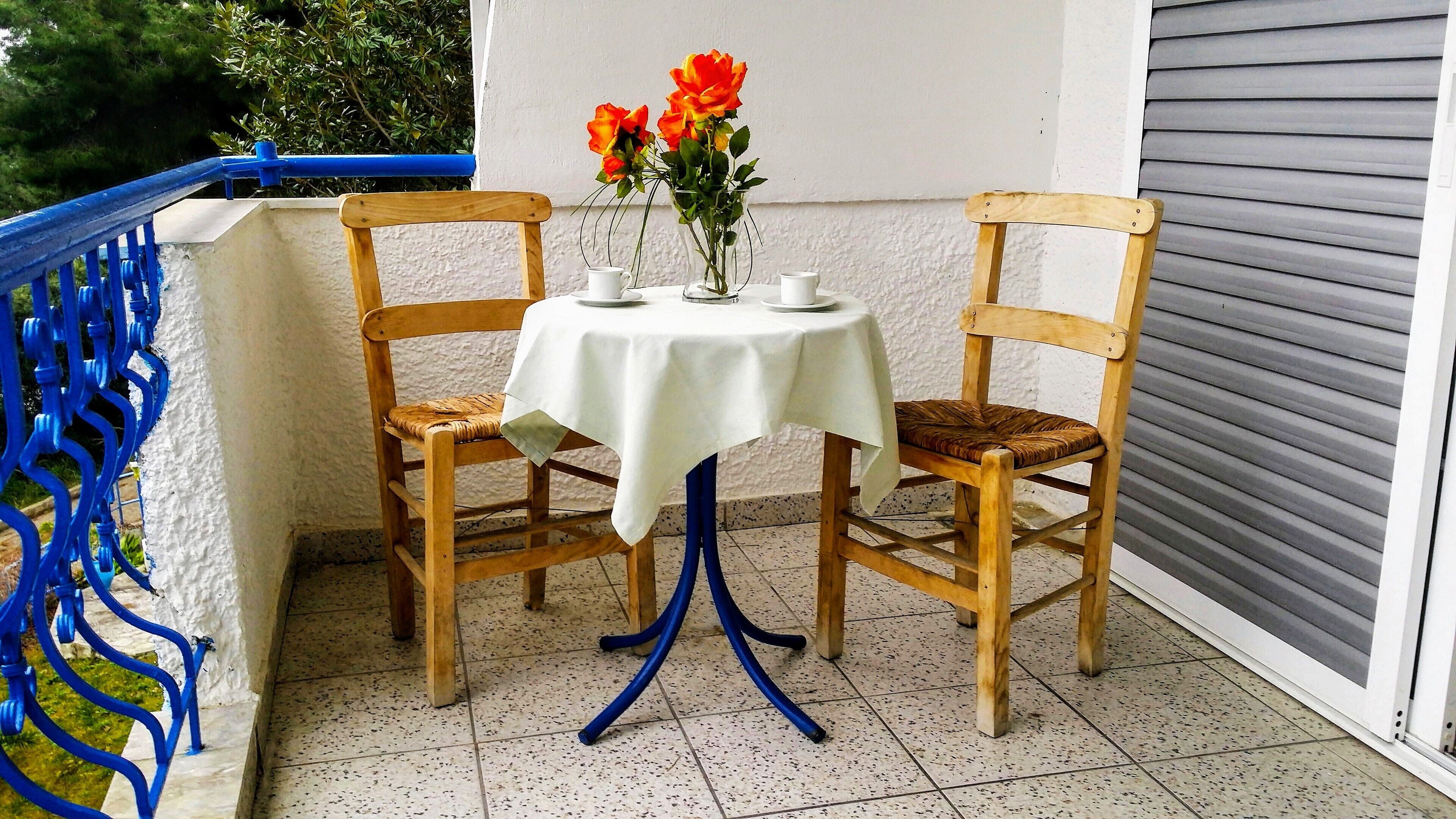Een kleine buitentafel met een wit tafelkleed, twee houten stoelen en een vaas met bloemen.
