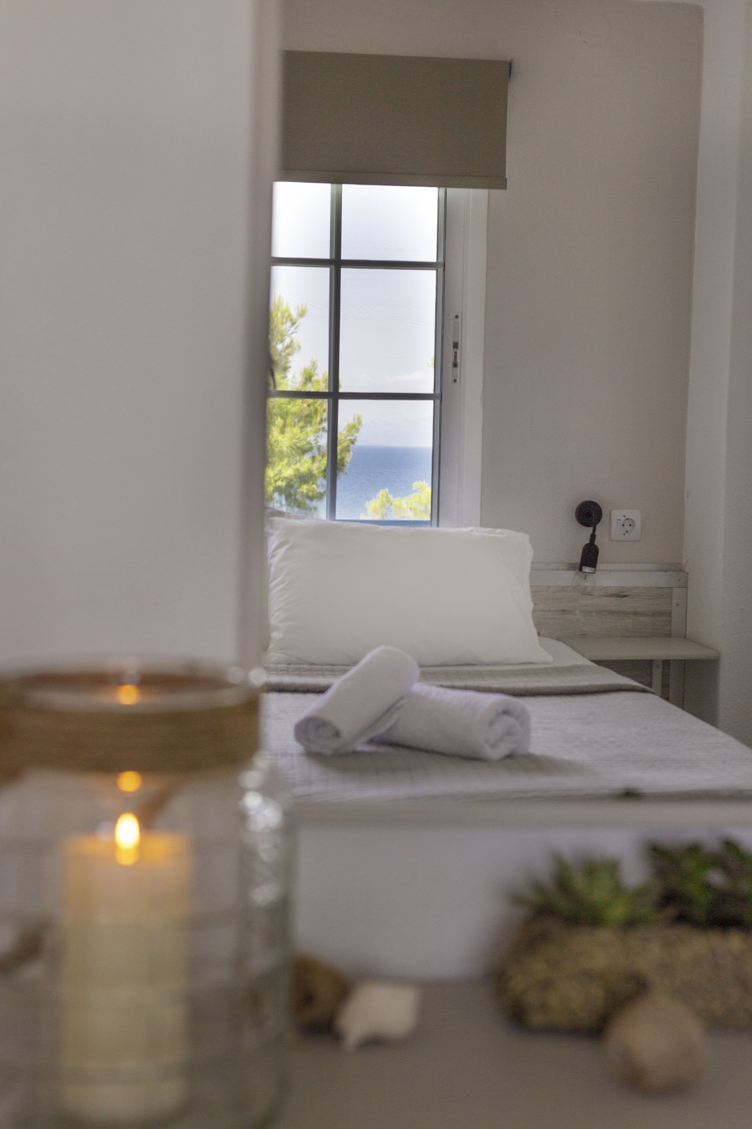 A bedroom with a bed, a nightstand, a lamp, a window with a view of the sea, and a candle on the bedside table.