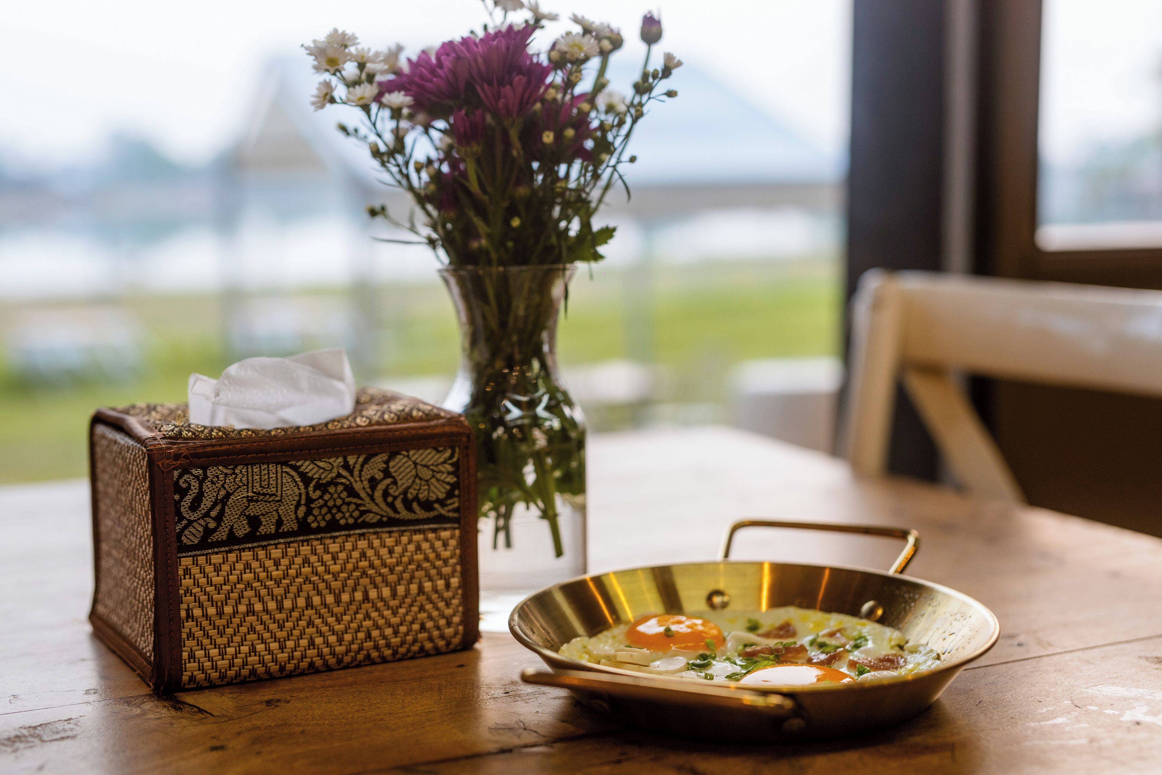 A wooden table with a tissue box, a vase with flowers, and a metal bowl with food.