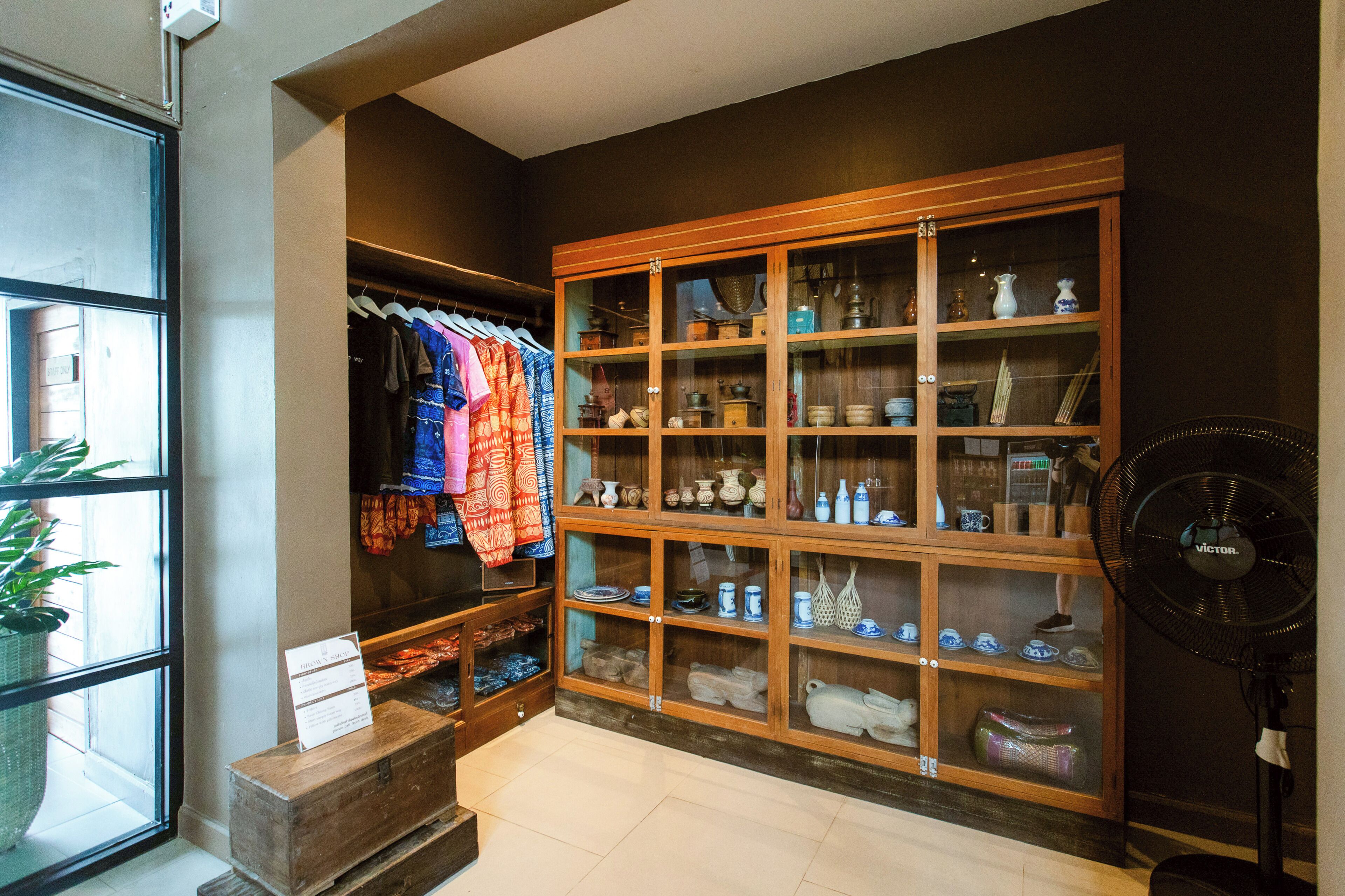 A room with a wooden cabinet displaying ceramics and textiles, a fan, and a window with a plant.