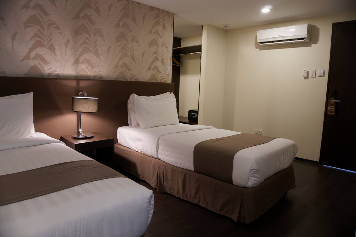 A hotel room with two beds, a bedside table with a lamp, an air conditioner, and a wardrobe.