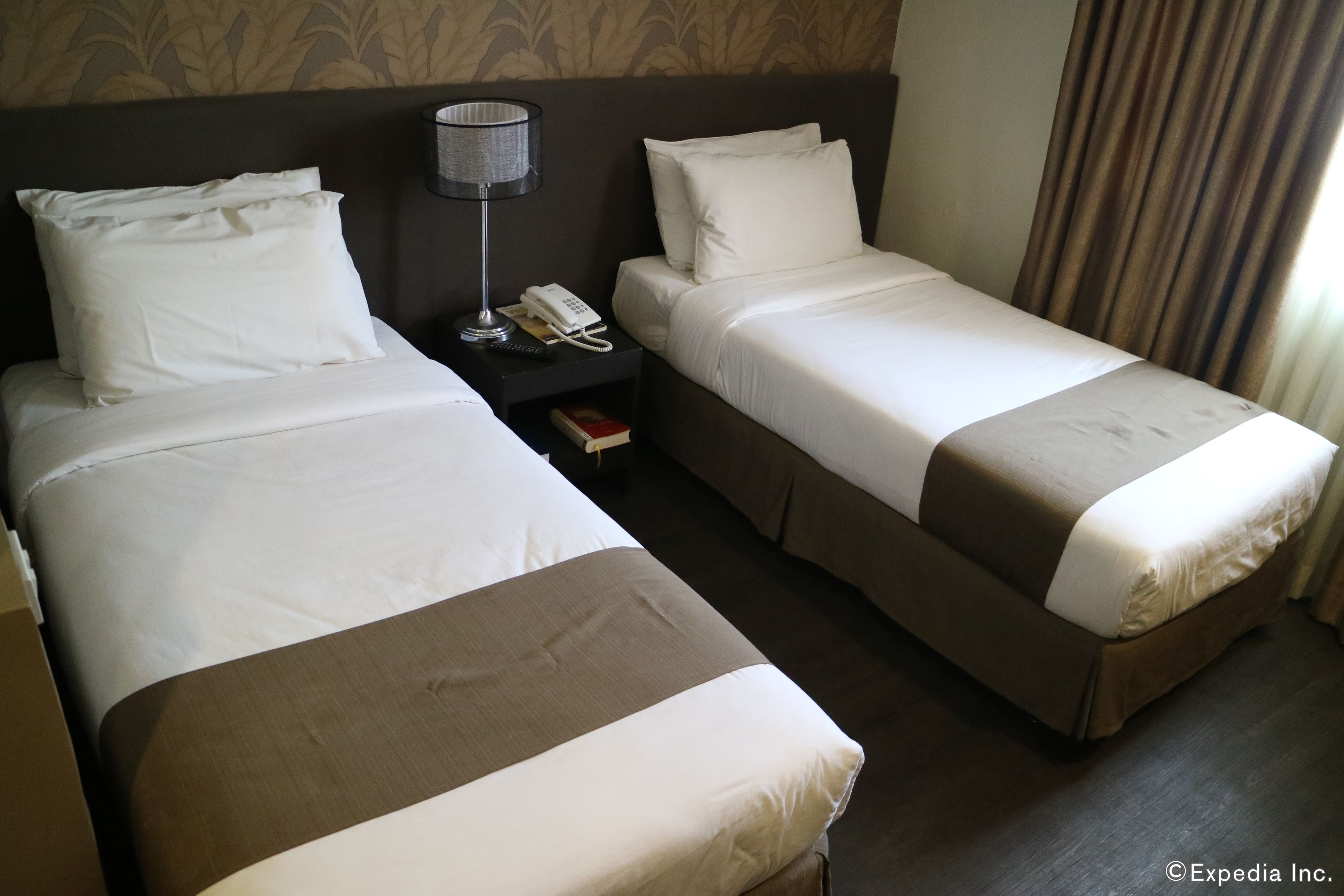 A hotel room with two single beds, a nightstand with a lamp, a phone, and a book.