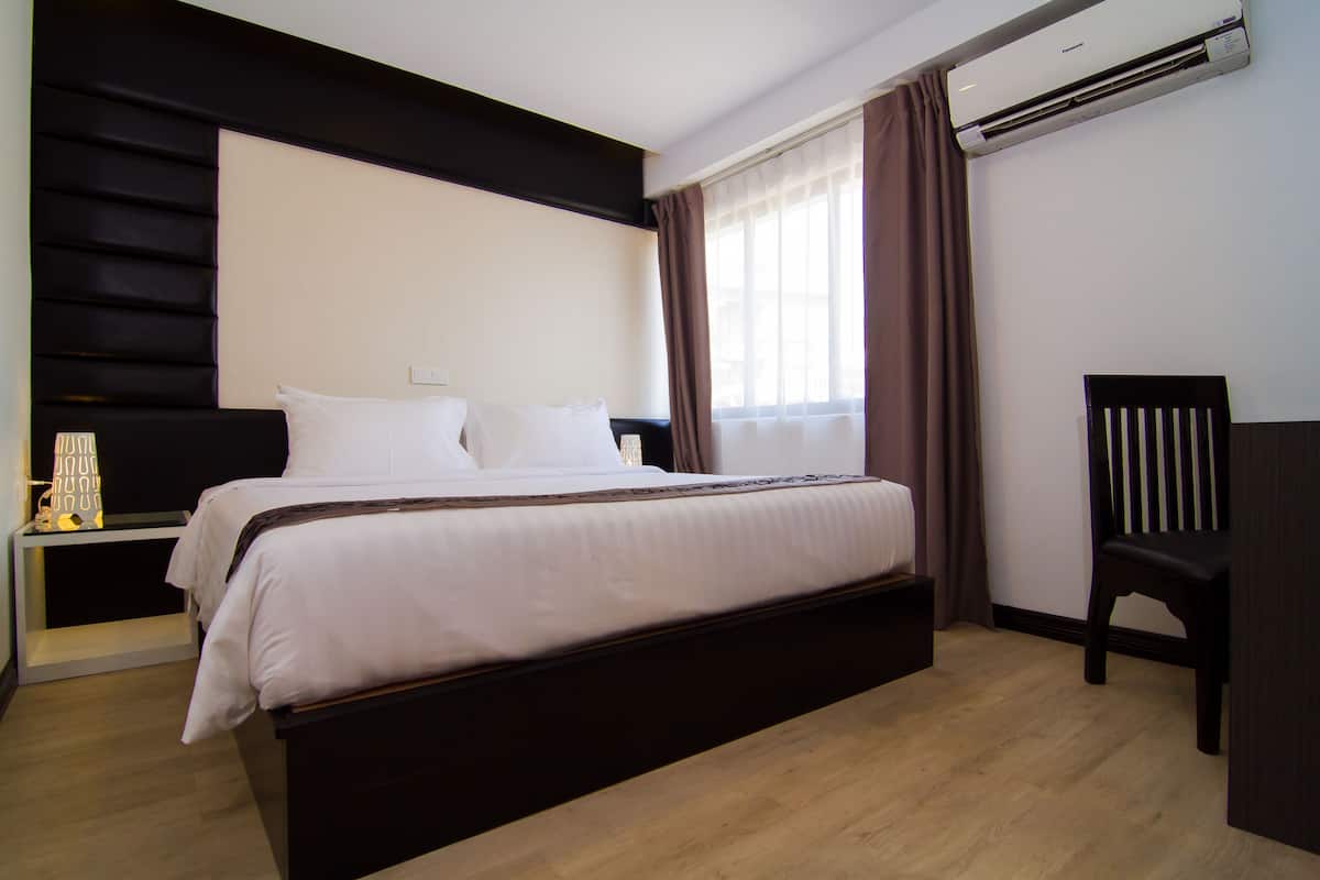A hotel room with a large bed, a bedside table, a chair, and an air conditioning unit.
