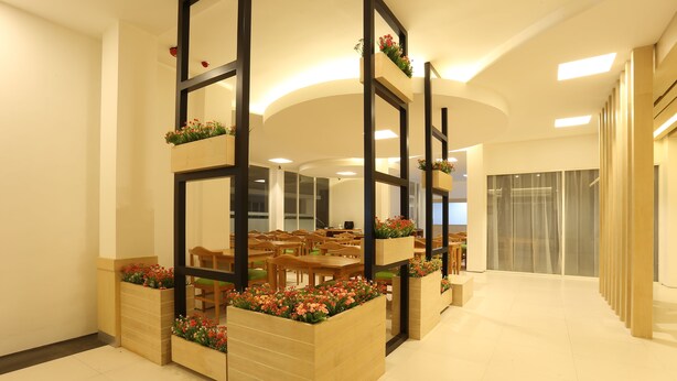 Eine moderne Hotelhalle mit Holzkübeln mit Blumen, einer Sitzecke mit Holztischen und -stühlen sowie einem großen Spiegel, der den Raum reflektiert.