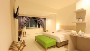 Ein Hotelzimmer mit einem Bett, einer grünen Bank, einem Holznachttisch, einem Regal mit Handtüchern und einem Fenster mit Vorhängen.