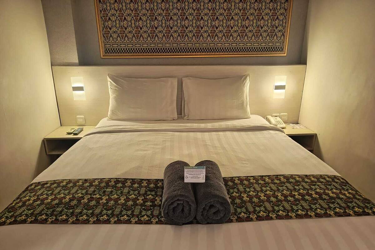 A neatly made bed with two pillows, a folded towel, and a patterned runner. There are bedside tables with lamps and a telephone on one side.