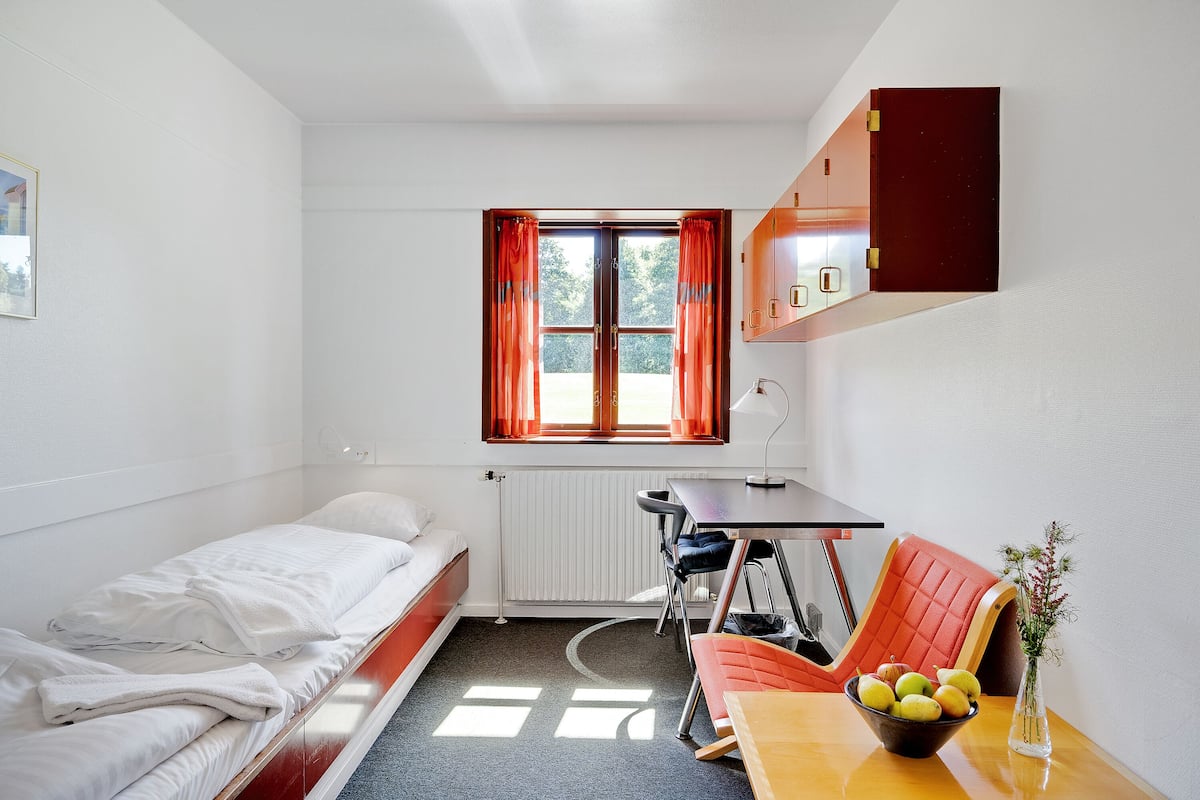 Una habitación pequeña y bien iluminada con una cama, un escritorio y una silla. Se ve una ventana con cortinas, un radiador y un jarrón con flores sobre el escritorio.