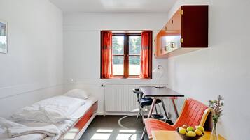 A small, well-lit room with a bed, a desk, and a chair. There is a window with curtains, a radiator, and a vase with flowers on the desk.